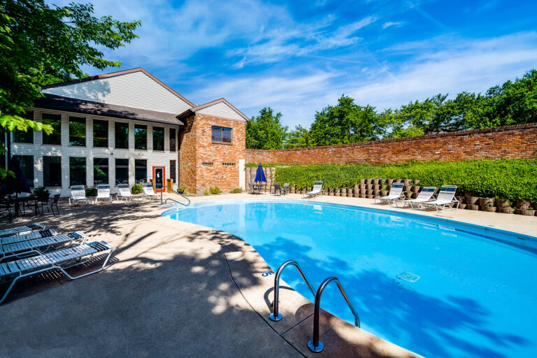 Swimming pool with chaise lounge seating, two story leasing office overlooking the pool deck, and tall brick wall on the far side with vines growing on it.
