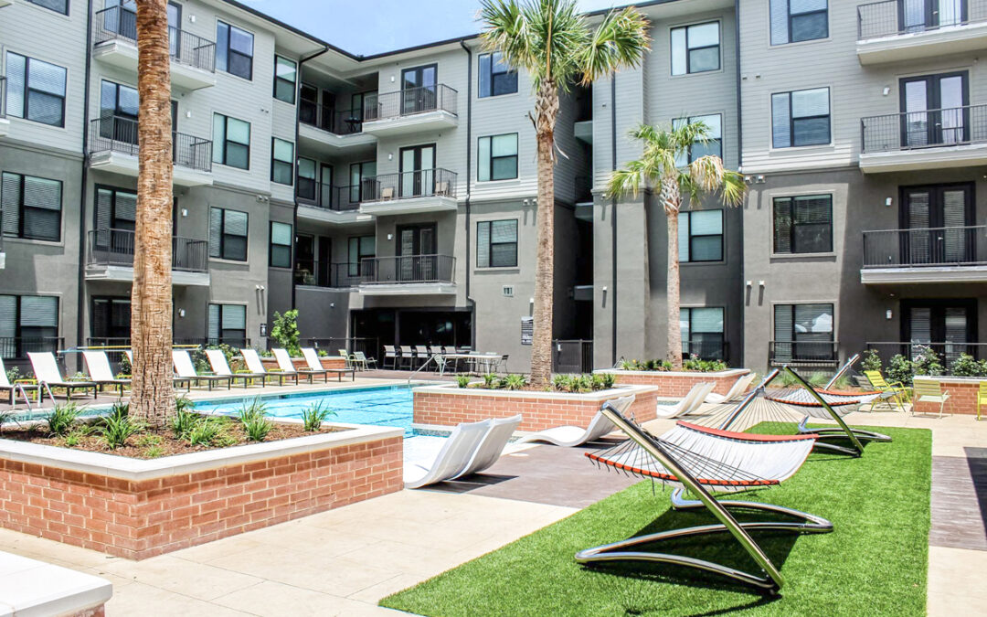 Pool with hammocks set between large resident building. Building is grey and four stories tall with palm trees.