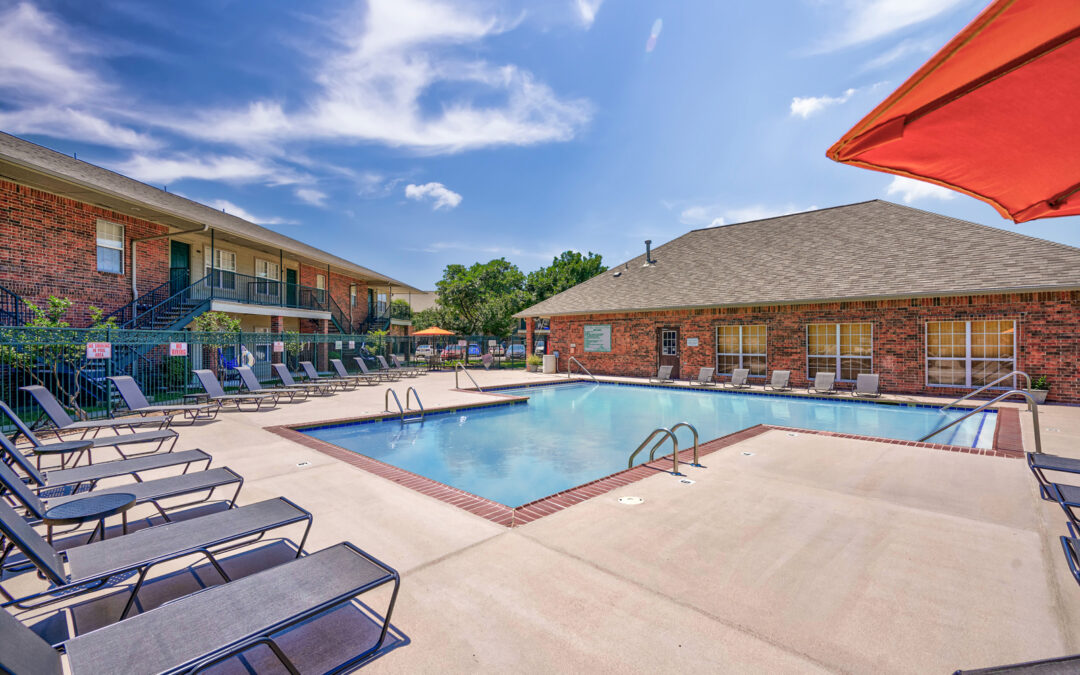 Community pool shown with chaise lounge seating and a red umbrella in the foreground. The back of the leasing office and a residential building are visible in the background.