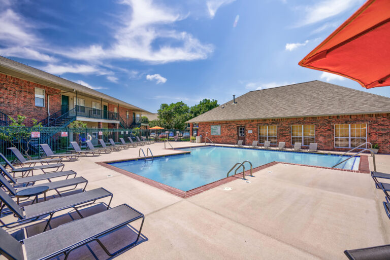 Community pool shown with chaise lounge seating and a red umbrella in the foreground. The back of the leasing office and a residential building are visible in the background.